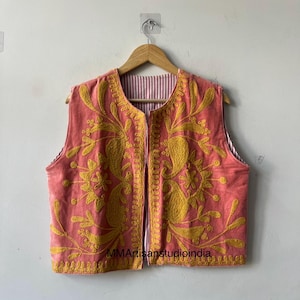 May include: A coral-coloured, sleeveless vest with detailed yellow floral embroidery. The vest has a striped lining and a full-length zip. It is displayed on a wooden hanger against a plain white wall. The text "MMArtisanstudioindia" is visible.