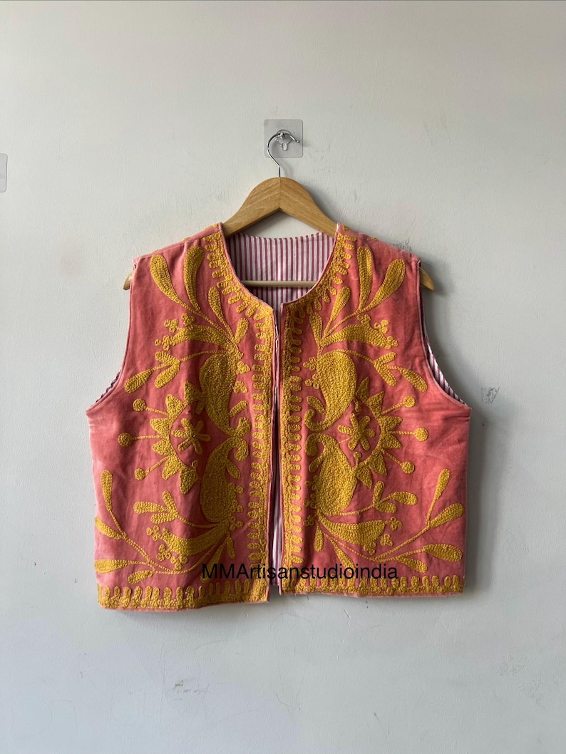 May include: A sleeveless, coral-colored vest with intricate yellow floral embroidery. The vest features a striped lining and a full-length zipper. The garment is displayed on a wooden hanger against a plain white wall. The text "MMArtisanstudioindia" is visible.