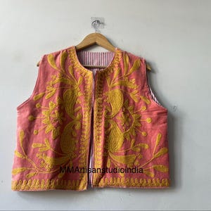 May include: A coral-colored vest with intricate yellow embroidery. The vest features a floral and paisley design, with a striped lining visible at the edges. The garment is displayed on a wooden hanger.