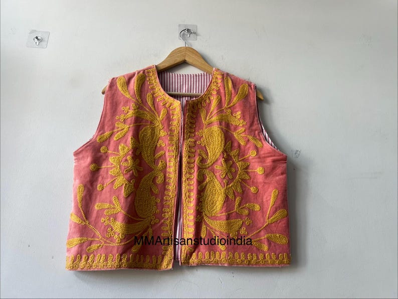May include: A coral-colored vest with intricate yellow embroidery. The vest features a floral and paisley design, with a striped lining visible at the edges. The garment is displayed on a wooden hanger.