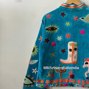 May include: A turquoise velvet jacket with colorful embroidered designs. The jacket features a variety of motifs, including stars, a cowboy boot, a cactus, and an eye. The collar has a striped rainbow design. The text "MMArtisanstudioindia" is visible.