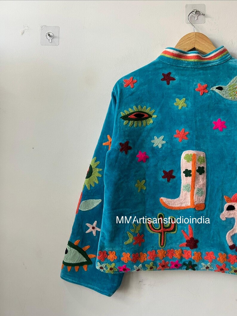 May include: A turquoise velvet jacket with colorful embroidered designs. The jacket features a variety of motifs, including stars, a cowboy boot, a cactus, and an eye. The collar has a striped rainbow design. The text "MMArtisanstudioindia" is visible.