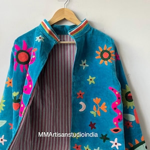 May include: Turquoise velvet jacket with colorful embroidered suns, flowers, stars, and a snake design. The jacket has a striped lining and a rainbow-striped collar. The text "MMArtisansstudioindia" is visible on the lining.