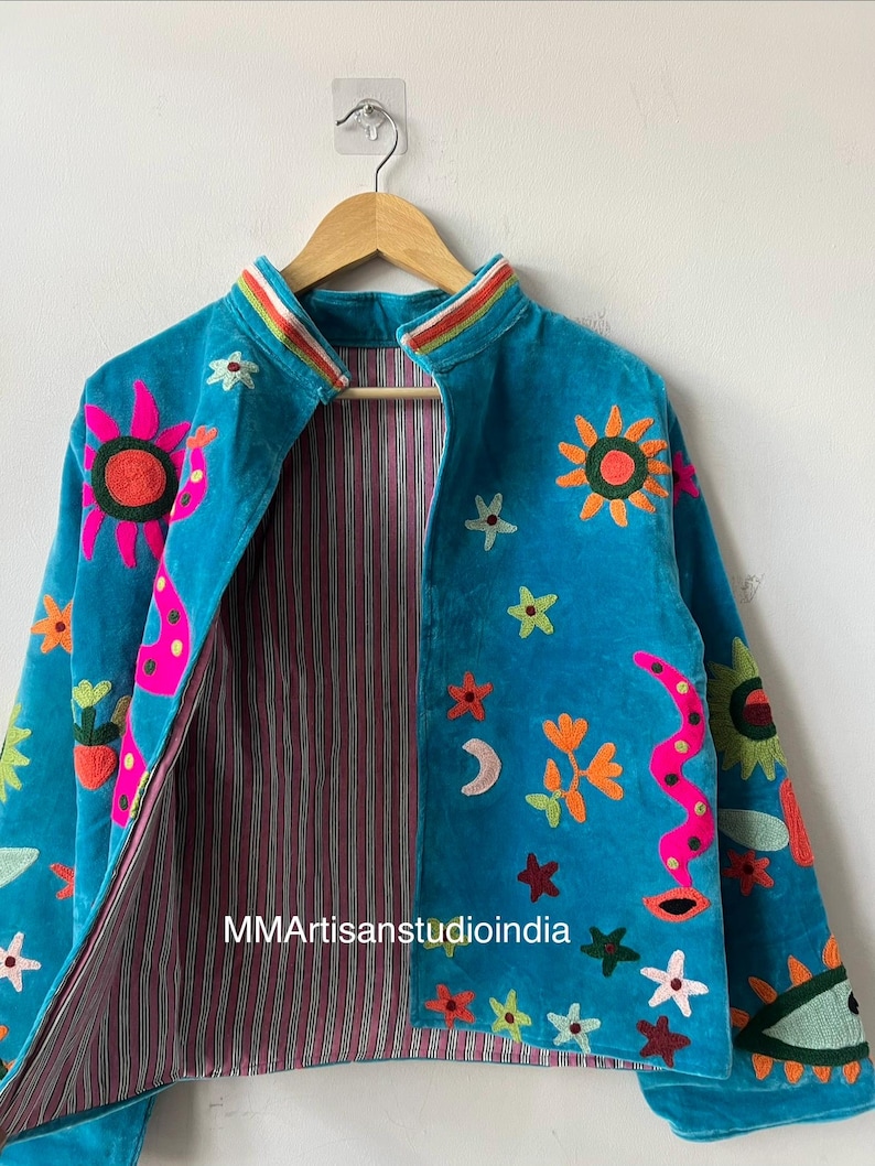 May include: Turquoise velvet jacket with colorful embroidered suns, flowers, stars, and a snake design. The jacket has a striped lining and a rainbow-striped collar. The text "MMArtisansstudioindia" is visible on the lining.