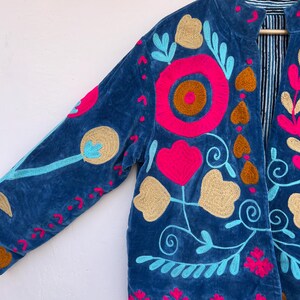 May include: A blue velvet jacket with vibrant floral embroidery in pink, gold, and turquoise. The jacket features a mandarin collar and is lined with a striped fabric. The embroidery includes flowers, hearts, and swirling vines.