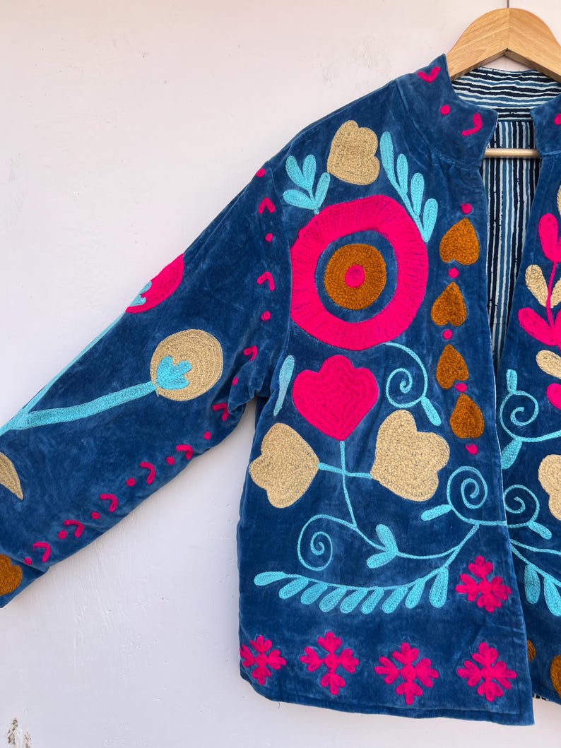 May include: A blue velvet jacket with vibrant floral embroidery in pink, gold, and turquoise. The jacket features a mandarin collar and is lined with a striped fabric. The embroidery includes flowers, hearts, and swirling vines.