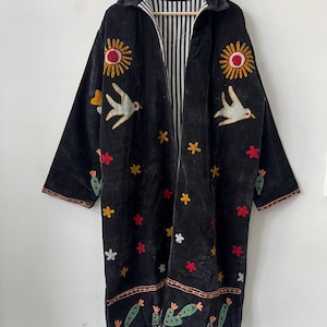 May include: Black velvet coat with embroidered suns, birds, flowers, and cacti. The coat has a striped lining and a collar. The embroidery features red, yellow, and white accents. The coat is hanging on a wooden hanger.