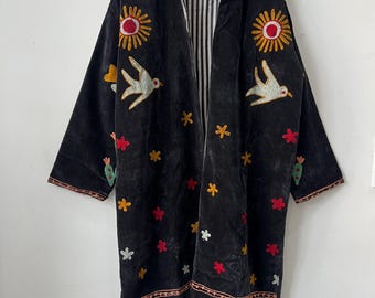 May include: Black velvet coat with embroidered suns, birds, flowers, and cacti. The coat has a striped lining and a collar. The embroidery features red, yellow, and white accents. The coat is hanging on a wooden hanger.