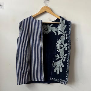 Puede incluir: Un chaleco sin mangas con la parte delantera a rayas y la espalda bordada con motivos florales. La parte delantera presenta rayas verticales azul marino y blancas. La espalda es azul marino con bordados florales en azul claro. El chaleco est&aacute; colgado de una percha de madera.