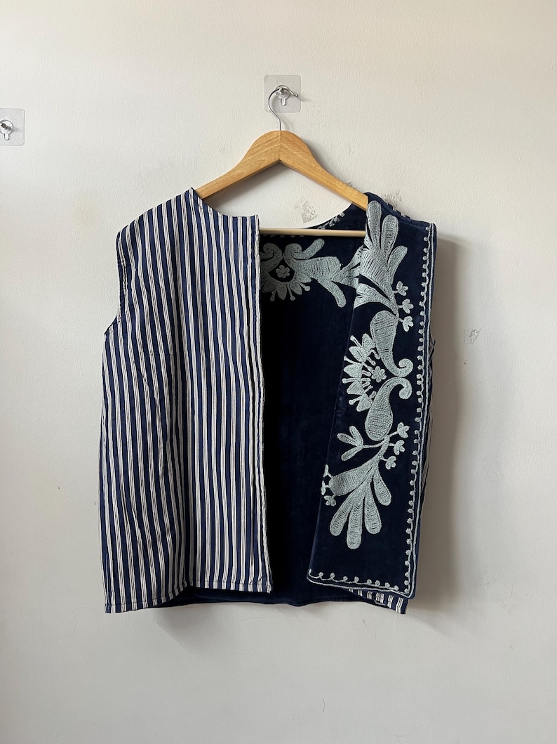 Puede incluir: Un chaleco sin mangas con la parte delantera a rayas y la espalda bordada con motivos florales. La parte delantera presenta rayas verticales azul marino y blancas. La espalda es azul marino con bordados florales en azul claro. El chaleco est&aacute; colgado de una percha de madera.