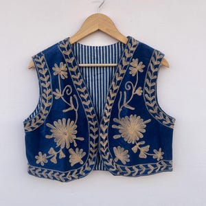 May include: A sleeveless, navy blue vest with intricate gold floral embroidery. The vest is hanging on a wooden hanger against a white wall. The interior lining features a striped pattern.