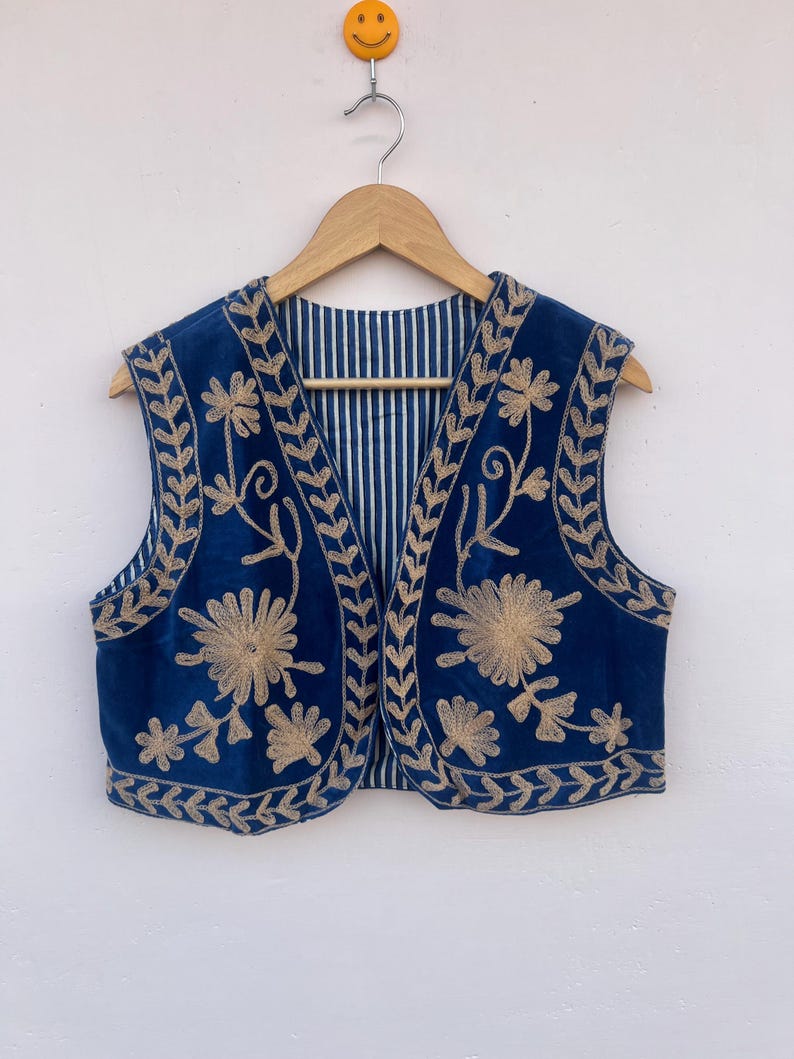 May include: A sleeveless, navy blue vest with intricate gold floral embroidery. The vest is hanging on a wooden hanger against a white wall. The interior lining features a striped pattern.