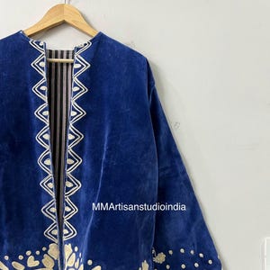 May include: A royal blue velvet jacket featuring cream-colored embroidered trim along the front opening and hem. The jacket has long sleeves and is displayed on a wooden hanger. The text MMArtisanstudioindia is visible on the fabric.