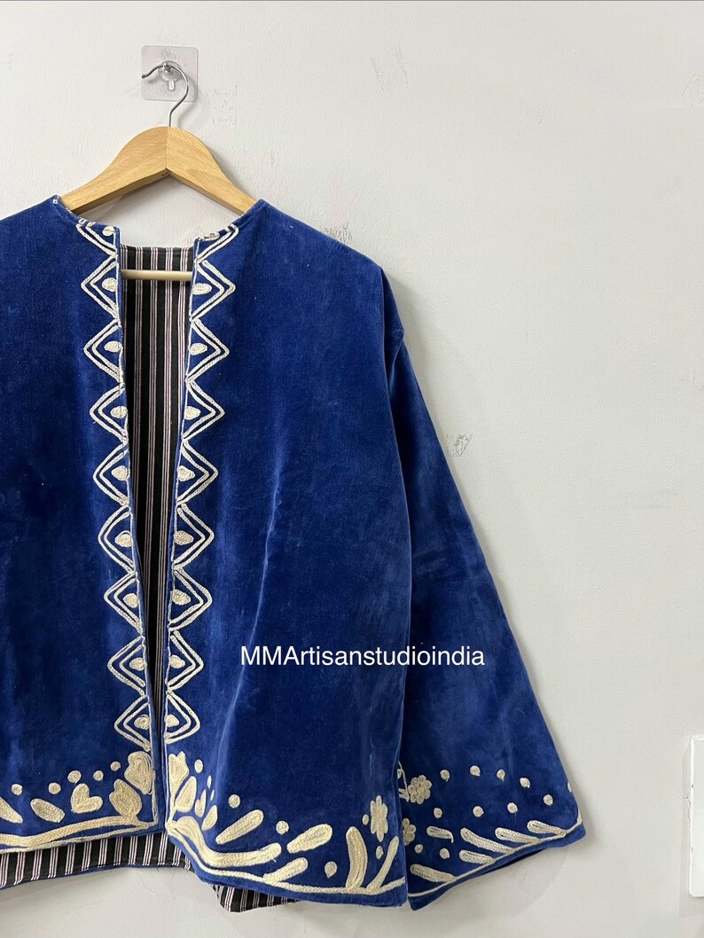 May include: A royal blue velvet jacket featuring cream-colored embroidered trim along the front opening and hem. The jacket has long sleeves and is displayed on a wooden hanger. The text MMArtisanstudioindia is visible on the fabric.