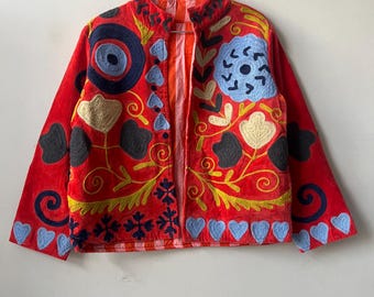 May include: A red jacket with colorful floral embroidery. The jacket features a stand-up collar and long sleeves. The embroidery includes blue, yellow, and white floral and heart-shaped designs. The jacket is hanging on a metal hanger.