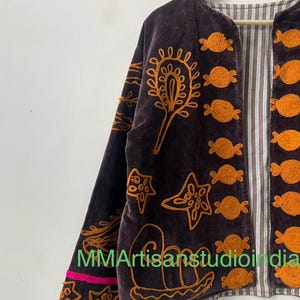 Puede incluir: Chaqueta de terciopelo marr&oacute;n oscuro con dise&ntilde;os bordados en naranja, incluyendo motivos florales y marinos. La chaqueta tiene un forro a rayas y un ribete rosa y naranja en la manga. El texto "MMArtisansstudioindia" es visible.