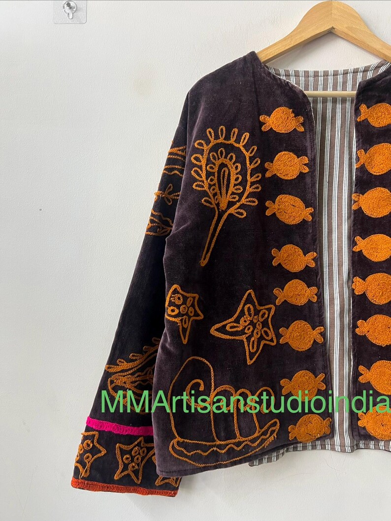 Puede incluir: Chaqueta de terciopelo marr&oacute;n oscuro con dise&ntilde;os bordados en naranja, incluyendo motivos florales y marinos. La chaqueta tiene un forro a rayas y un ribete rosa y naranja en la manga. El texto "MMArtisansstudioindia" es visible.