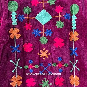May include: Close-up of a magenta textile with colorful, embroidered floral and geometric designs. The embroidery includes orange, pink, green, and blue flowers, along with a turquoise diamond shape. The text "MMArtisanstudioindia" is visible.