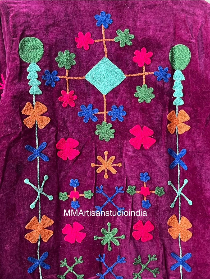 May include: Close-up of a magenta textile with colorful, embroidered floral and geometric designs. The embroidery includes orange, pink, green, and blue flowers, along with a turquoise diamond shape. The text "MMArtisanstudioindia" is visible.