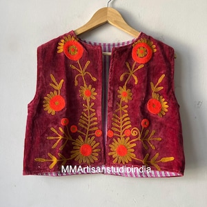 Puede incluir: Chaleco burdeos sin mangas con bordados florales en naranja y dorado. El chaleco est&aacute; colgado de una percha de madera. El texto "MMArtisanstudioindia" es visible en la parte inferior. Fondo blanco.