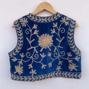 May include: A sleeveless, blue vest with intricate gold floral embroidery. The vest features a round neckline and a cropped length. The embroidery includes floral and leaf patterns, creating a detailed and ornate design. The vest is hanging on a wooden hanger.