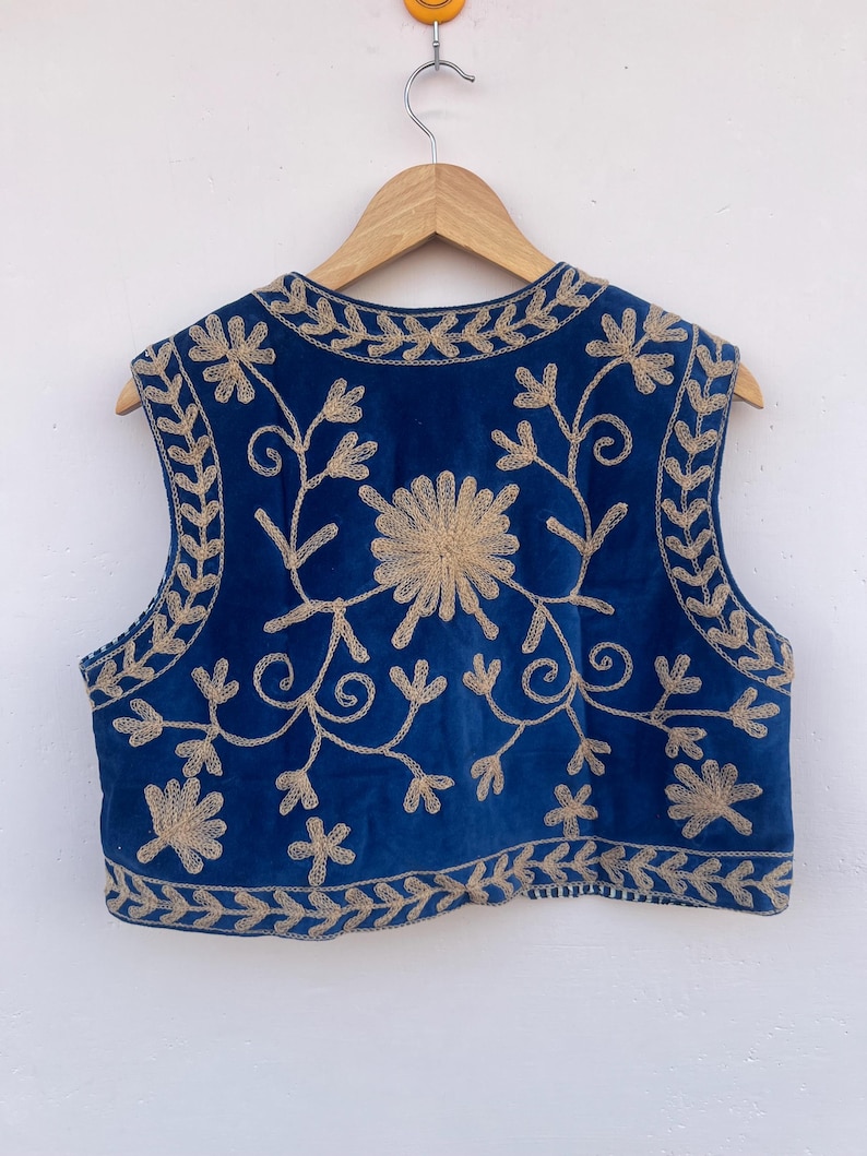 May include: A sleeveless, blue vest with intricate gold floral embroidery. The vest features a round neckline and a cropped length. The embroidery includes floral and leaf patterns, creating a detailed and ornate design. The vest is hanging on a wooden hanger.