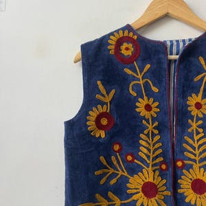 May include: A sleeveless, navy blue vest with embroidered floral designs in yellow and red. The vest has a striped lining and is hanging on a wooden hanger. The text "MMArtisanstudioindia" is visible at the bottom.