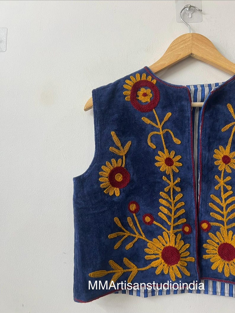May include: A sleeveless, navy blue vest with embroidered floral designs in yellow and red. The vest has a striped lining and is hanging on a wooden hanger. The text "MMArtisanstudioindia" is visible at the bottom.