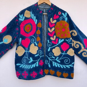 May include: A blue velvet jacket with colorful floral embroidery. The jacket features a round collar and long sleeves. The embroidery includes pink, gold, and turquoise accents, with a striped lining visible at the open front.