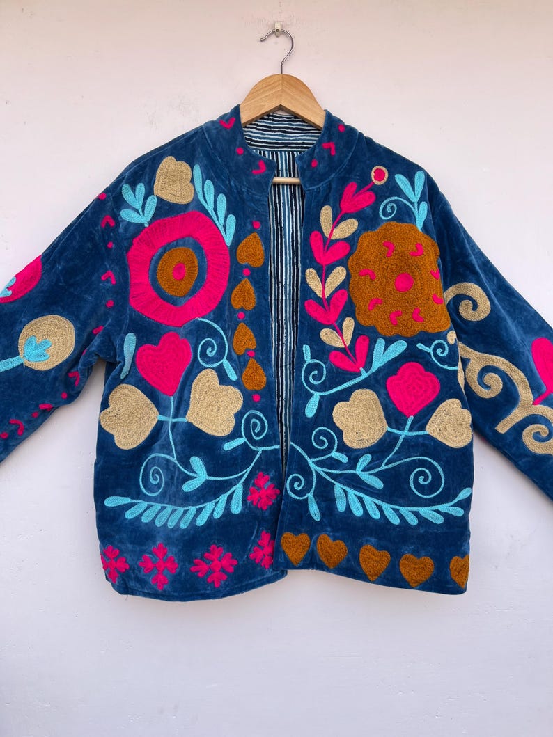 May include: A blue velvet jacket with colorful floral embroidery. The jacket features a round collar and long sleeves. The embroidery includes pink, gold, and turquoise accents, with a striped lining visible at the open front.