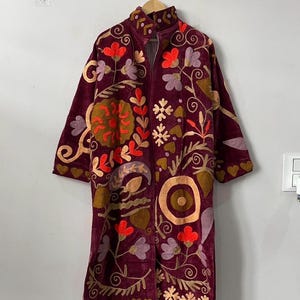 Könnte beinhalten: Ein knielanger Mantel mit langen Ärmeln aus tiefburgunderfarbenem Samt, verziert mit aufwendigen, mehrfarbigen Blumen- und abstrakten Stickereien in Orange-, Lavendel- und Goldtönen. Das Kleidungsstück hat einen Stehkragen und den Text MMArtisanstudioindia am Saum.