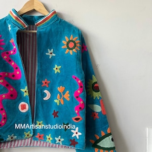 May include: Turquoise velvet jacket with colorful embroidered celestial and floral designs. Features a pink and orange snake-like pattern, suns, stars, and an eye. The jacket is lined with a striped fabric and hangs on a wooden hanger.
