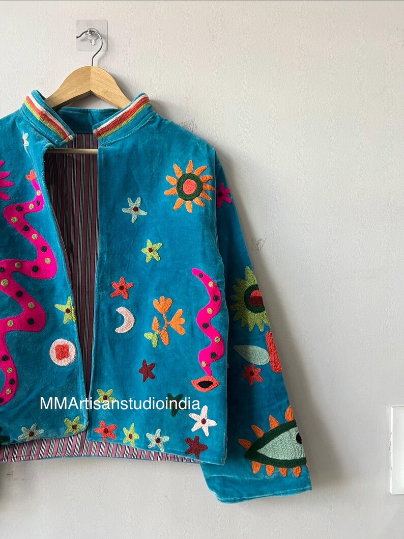 May include: Turquoise velvet jacket with colorful embroidered celestial and floral designs. Features a pink and orange snake-like pattern, suns, stars, and an eye. The jacket is lined with a striped fabric and hangs on a wooden hanger.