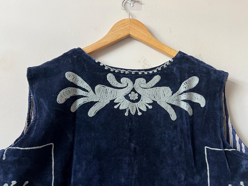 Puede incluir: Una prenda sin mangas de color azul marino con dise&ntilde;os florales bordados en azul claro. La prenda est&aacute; colgada de una percha de madera. El bordado est&aacute; centrado en el pecho y a lo largo del escote.