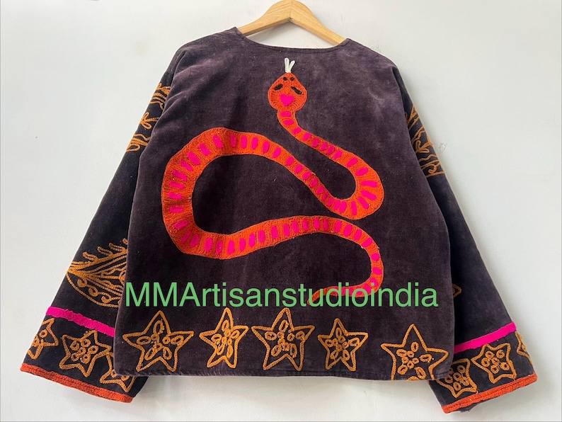 Puede incluir: Chaqueta marr&oacute;n oscuro con un gran dise&ntilde;o de serpiente bordada en naranja y rosa en la espalda. La chaqueta tiene bordados de estrellas y flores naranjas en la parte inferior y en las mangas. El texto "MMArtisanstudioindia" es visible.