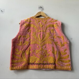 May include: A sleeveless, pink vest with intricate gold embroidery. The embroidery features floral and leaf patterns. The vest is hanging on a wooden hanger against a white wall. The text "MMArtisanstudioindia" is visible.