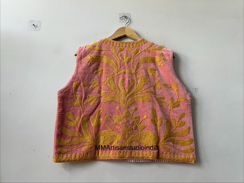 May include: A sleeveless, pink vest with intricate gold embroidery. The embroidery features floral and leaf patterns. The vest is hanging on a wooden hanger against a white wall. The text "MMArtisanstudioindia" is visible.