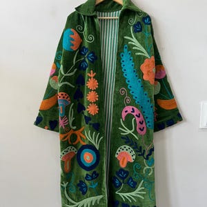 Può includere: Un cappotto lungo aperto in velluto verde, caratterizzato da vivaci ricami multicolori di fiori, funghi e forme astratte. La fodera interna è visibile e presenta un motivo a righe verticali bianche e verdi.