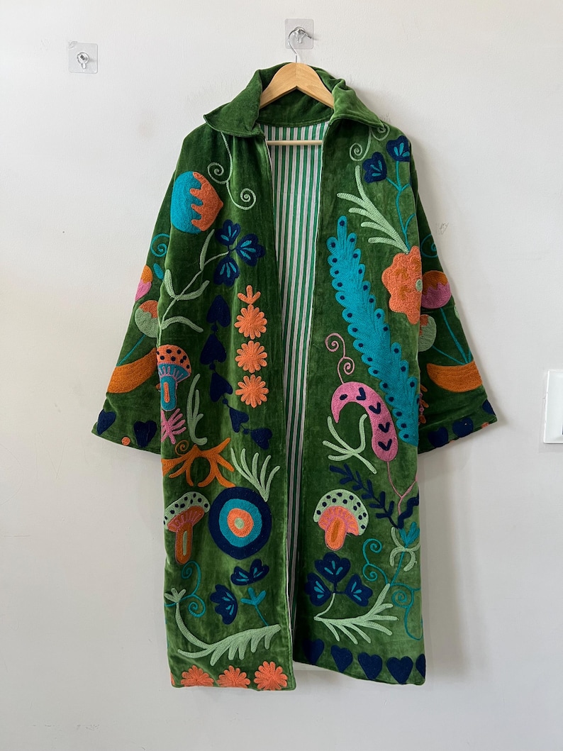 May include: A long, open-front coat made of green velvet, featuring vibrant, multi-colored embroidery of flowers, mushrooms, and abstract shapes. The interior lining is visible, displaying a white and green vertical striped pattern.