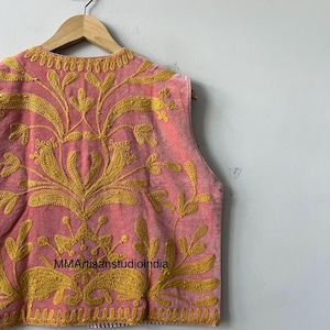 May include: A sleeveless pink vest with intricate gold embroidery featuring floral and leaf patterns. The vest is hanging on a wooden hanger against a plain white wall. The text "MMArtisanstudioindia" is visible on the vest.