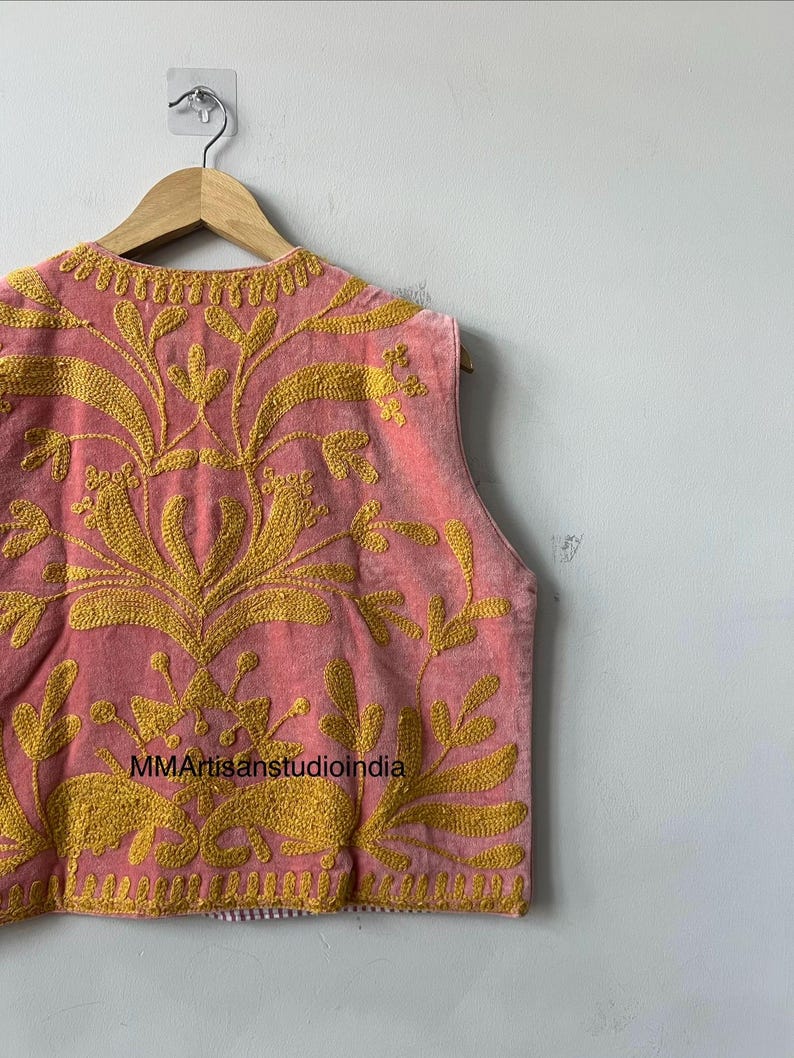 May include: A sleeveless pink vest with intricate gold embroidery featuring floral and leaf patterns. The vest is hanging on a wooden hanger against a plain white wall. The text "MMArtisanstudioindia" is visible on the vest.