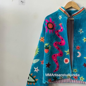 May include: A turquoise velvet jacket with colorful embroidered celestial and floral designs. The jacket features a pink sun, a pink snake, stars, and a crescent moon. The lining has a striped pattern. The text "MMArtisanstudioindia" is visible.