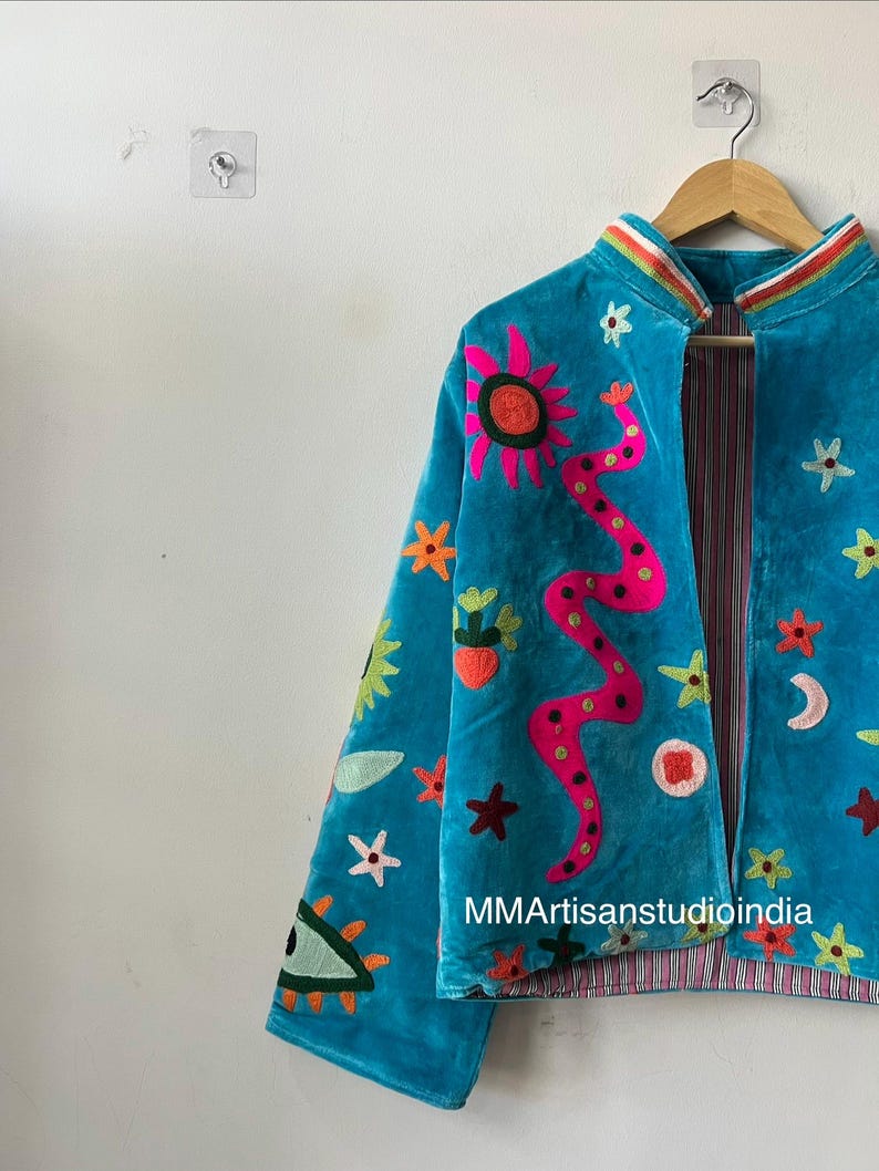 May include: A turquoise velvet jacket with colorful embroidered celestial and floral designs. The jacket features a pink sun, a pink snake, stars, and a crescent moon. The lining has a striped pattern. The text "MMArtisanstudioindia" is visible.