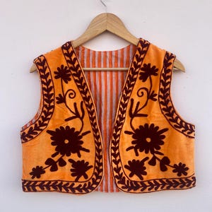 May include: Orange vest with dark brown floral embroidery and trim. The vest has a striped orange and white lining. The vest is hanging on a wooden hanger.
