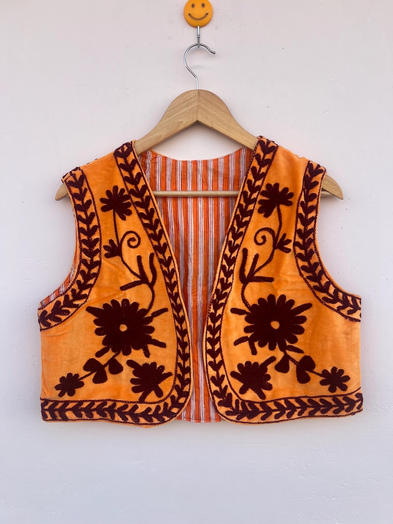 May include: Orange vest with dark brown floral embroidery and trim. The vest has a striped orange and white lining. The vest is hanging on a wooden hanger.