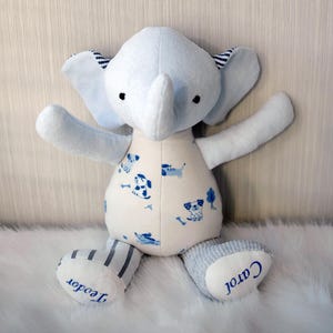 Könnte beinhalten: Ein hellblaues Plüsch-Elefanten-Spielzeug mit weißem Körper und blauen Akzenten. Der Elefant hat große Ohren, einen Rüssel und einen gemusterten Bauch mit blauen Hunde-Illustrationen. Die Füße sind grau und weiß gestreift, mit den Namen "Teodor" und "Carol" blau gestickt.