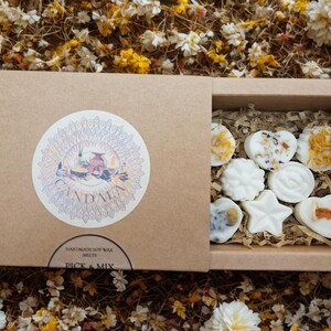 May include: A brown cardboard box with a sliding lid, featuring a circular sticker with the word "Candala" and floral illustrations. Inside, various white soy wax melts in different shapes are visible. The box is surrounded by dried flowers.