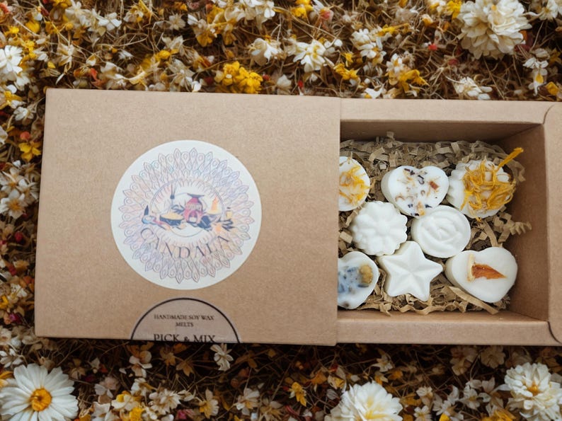 May include: A brown cardboard box with a sliding lid, featuring a circular sticker with the word "Candala" and floral illustrations. Inside, various white soy wax melts in different shapes are visible. The box is surrounded by dried flowers.