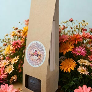 May include: A light brown cardboard box with a decorative label and a small window, containing incense sticks. The box is set against a backdrop of colourful flowers in shades of pink, orange, yellow, and white.