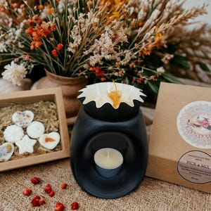 May include: A black essential oil wax melt warmer with a lit tealight candle inside. A small wooden box contains wax melts in various shapes. A brown box with a circular sticker is next to the warmer. Dried flowers are in the background.